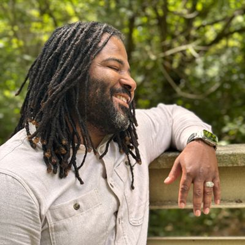 Man smiling outdoors near greenery.